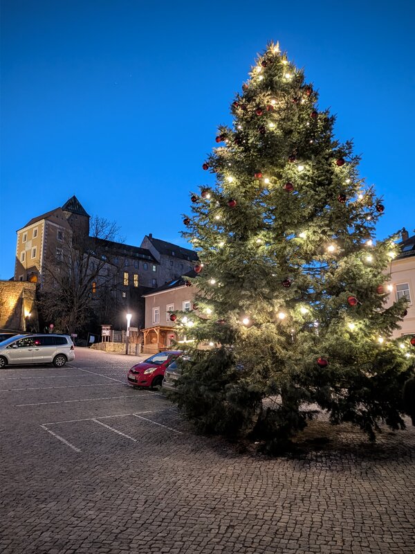 Großer geschmückter Weihnachtsbaum mit Lichterkette und roten Kugeln auf einem gepflasterten Platz, historische Gebäude im Hintergrund bei Abenddämmerung.