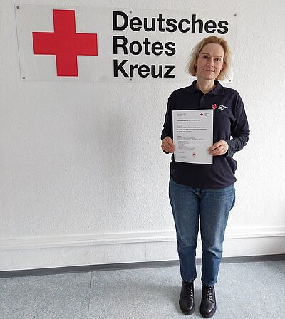 Vom Flüchtling zur Kriseninterventionshelferin Frau Yuliia Nahorna steht in blauen Jeans, dunklen Chelsea Boots und dunklem Pullover lächelnd vor einem großen Schild des Deutschen Roten Kreuzes und hält ihre Teilnahmebescheinigung in die Kamera.