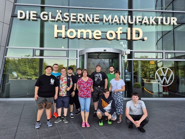 Auf dem Bild zu sehen ist eine Gruppe von jungen Leuten, die vor dem Eingang der Gläsernen Manufaktur in Dresden posieren, die aus einer Glaswand und einem goldenen Schriftzug besteht.