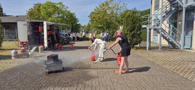 Zwei Frauen stehen auf einem gepflasterten Platz und löschen mit Feuerlöschern einen kleinen Brand in einer rechteckigen Metallwanne. Im Hintergrund beobachten mehrere Personen die Szene, einige sitzen auf Bänken, andere stehen. Rechts ist ein Treppenaufgang an einem Gebäude zu sehen, links stehen Gasflaschen und weitere Ausrüstungsgegenstände. Die Sonne scheint, Bäume spenden teilweise Schatten.