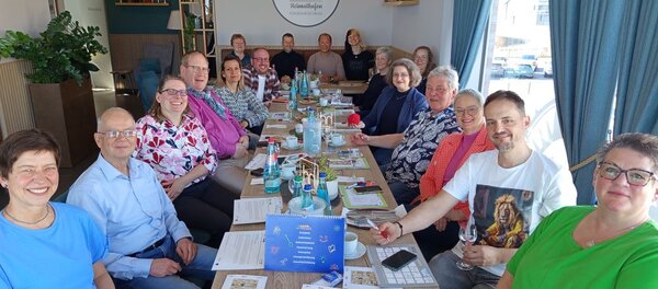 Eine Gruppe von 16 lächelnden Erwachsenen sitzt gemeinsam an einem langen, gedeckten Tisch in einem hellen Raum mit großen Fenstern. Die Personen wirken entspannt und fröhlich, einige halten Gläser oder Tassen in der Hand. Auf dem Tisch sind verschiedene Unterlagen, ein Kalender, Stifte und kleine Dekorationen zu sehen. Im Hintergrund ist ein Schild mit der Aufschrift 'Heimathafen' zu erkennen