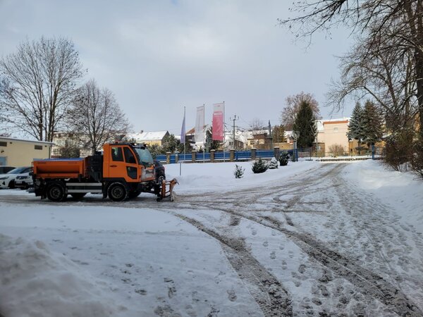 Ein orangefarbener Schneeräum-Lkw des Bauhofs Hohenstein-Ernstthal steht auf einer verschneiten Zufahrt vor dem Gelände der Euro-Schulen Hohenstein-Ernstthal. Der Lkw ist mit einem Schneepflug ausgestattet und befindet sich auf einer geräumten Fahrspur, die von zahlreichen Fußspuren und Fahrspuren durchzogen ist. Im Hintergrund sind mehrere Fahnenmasten mit Fahnen, ein blauer Zaun, einige Gebäude und winterliche Bäume zu sehen. Die Szene zeigt eine typische Winterlandschaft mit klarem Himmel und Schnee auf dem Boden.