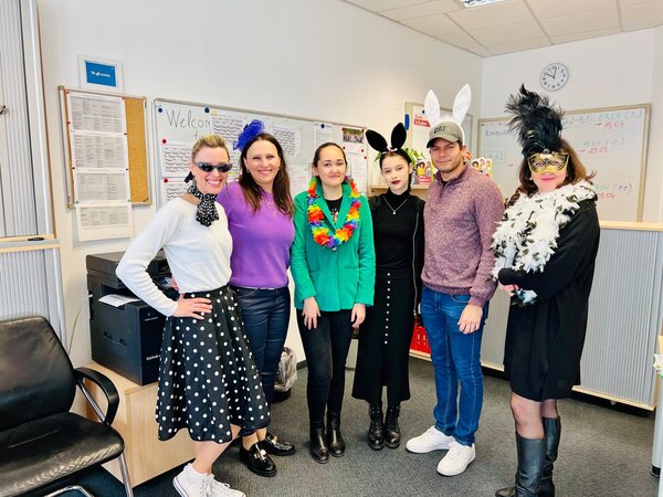 Eine Gruppe von sechs Personen in bunten, kreativen Kostümen posiert in einem Büro vor einem Whiteboard mit verschiedenen Notizen. Einige tragen Hasenohren, eine Person hat eine Federboa und eine Maske, während eine andere im Rockabilly-Stil mit Sonnenbrille und gepunktetem Rock gekleidet ist.