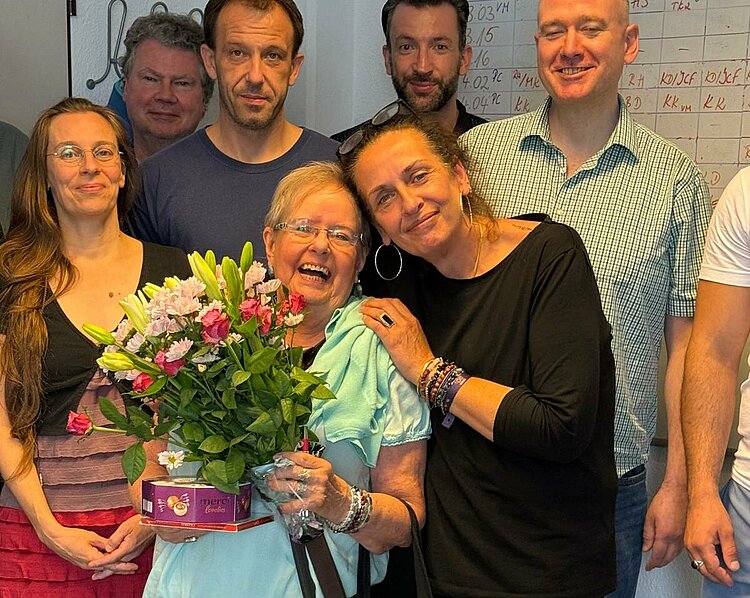 Bruni Drewitz – Die Geheimwaffe der Erwachsenenbildung Eine fröhliche Gruppe von Menschen posiert in einem Raum vor einem Whiteboard. Im Vordergrund steht eine ältere Frau mit Blumenstrauß und einer Schachtel Pralinen, sie wird herzlich von einer anderen Frau umarmt. Die Gruppe lächelt in die Kamera.