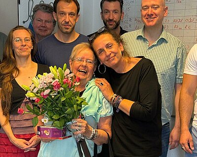 Bruni Drewitz – Die Geheimwaffe der Erwachsenenbildung Eine fröhliche Gruppe von Menschen posiert in einem Raum vor einem Whiteboard. Im Vordergrund steht eine ältere Frau mit Blumenstrauß und einer Schachtel Pralinen, sie wird herzlich von einer anderen Frau umarmt. Die Gruppe lächelt in die Kamera.