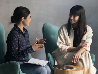 Zwei Frauen sitzen sich in grünen Sesseln gegenüber und führen ein Gespräch. Die Frau auf der linken Seite trägt ein dunkelblaues, gepunktetes Hemd und Jeans und hält einen Ordner auf ihrem Schoß. Die Frau auf der rechten Seite trägt eine cremefarbene Jacke und ein langes, cremefarbenes Kleid und hat ein Tablet auf einem kleinen Holztisch neben sich. Beide wirken entspannt und engagiert im Gespräch.