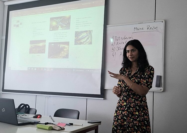 Ein junger weibliche Teilnehmerin steht, in einem sommerlichen Kleid, vorn am Whiteboard im Unterrichtsraum und hält ihre Präsentation vor ihren Mitteilnehmenden des B2-Sprachkurses an den Euro-Schulen Magdeburg.