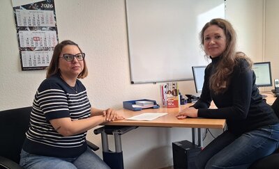 Die Teilnehmende und ihre Coachin sitzen sich an einem Schreibtisch gegenüber und lächeln in die Kamera. Beide tragen Brillen und haben schulterlanges Haar. Die Frau links trägt ein gestreiftes Oberteil, die Frau rechts ein einfarbiges, dunkles Oberteil. Auf dem Tisch liegen einige Blätter Papier. Im Hintergrund hängen zwei Wandkalender und ein Whiteboard an der Wand. Auf dem Schreibtisch stehen ein Aktenordner, ein Prospektständer mit buntem Flyer sowie weitere Büromaterialien.
