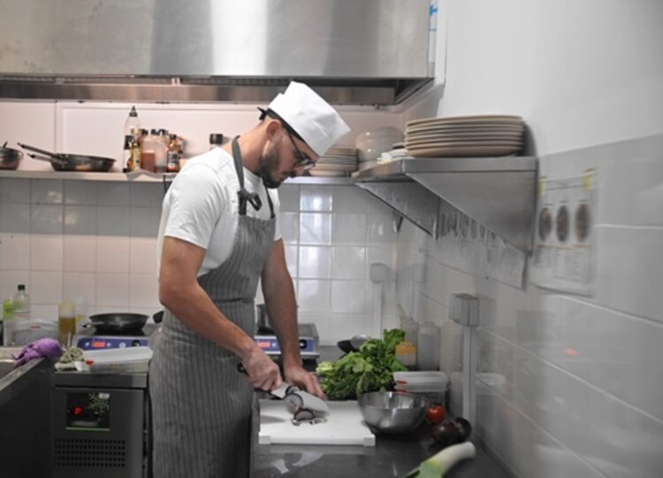 Ein junger Mann mit Kochschürze und Kochmütze steht in der Küche und schneidet mit einem Messer Gemüse in Scheiben.