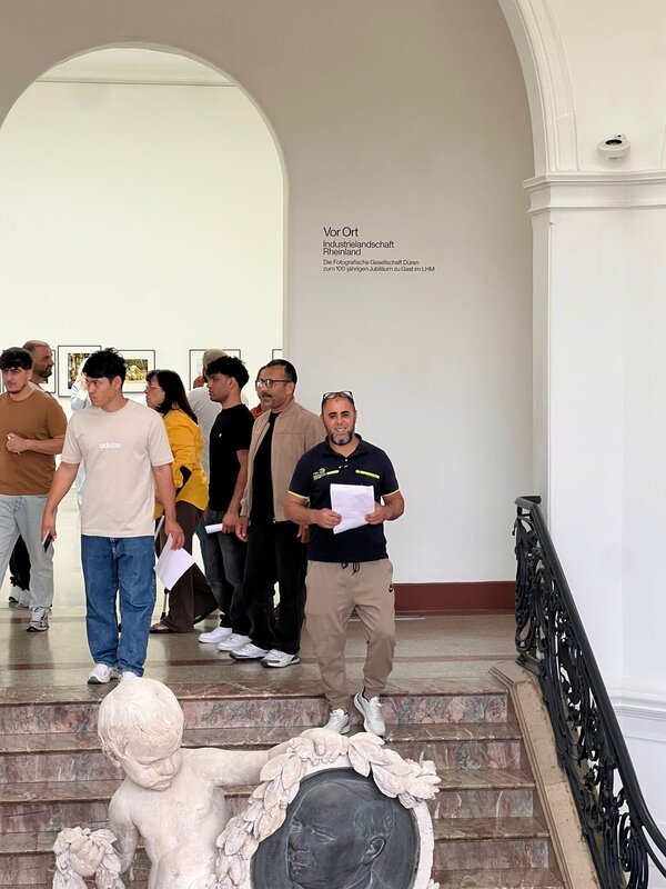 Gruppe von Teilnehmenden im Museum am oberen Treppenabsatz unter einem großen Rundbogen; mehrere Personen halten Unterlagen in der Hand, im Vordergrund ist die Steinfigur am Geländer zu sehen.