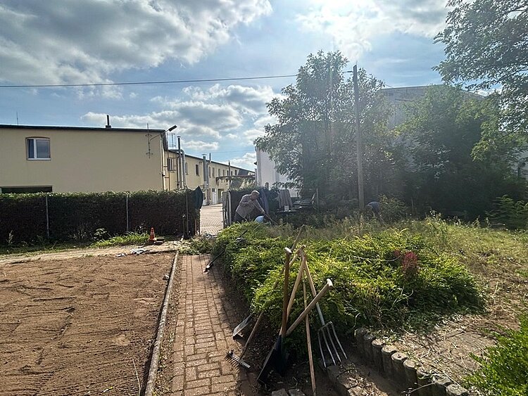 Blick auf den Garten, mit Häusern im Hintergrund und einem Teilnehmenden, der in einem Beet mit einem Spaten arbeitet. Es sind Spaten und Harken zu sehen und links ein umgepflügtes Feld.