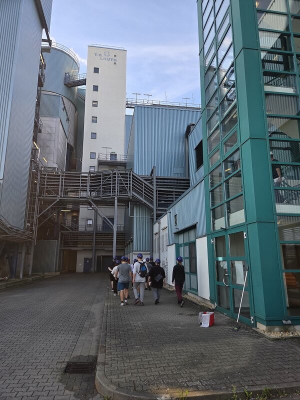 Mehrere Auszubildende mit Schutzhelmen gehen durch das Gelände einer Industrieanlage. Umgeben sind sie von großen, blau-grauen Produktionsgebäuden, Rohrleitungen, Metalltreppen und Laufstegen. Rechts befindet sich ein verglastes Gebäude mit türkisfarbenem Rahmen.