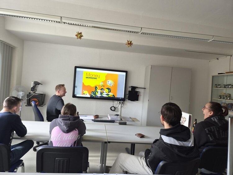 Eine Gruppe junger Auszubildender sitzt um einen Konferenztisch in einem Besprechungsraum und verfolgt aufmerksam eine Präsentation auf einem großen Bildschirm. Die Präsentationsfolie mit dem Titel „Geldmarketing“ ist mit einer farbenfrohen Illustration versehen. Der Raum ist hell erleuchtet, und im Hintergrund sind verschiedene Gegenstände wie ein Ventilator, ein Telefon und Regale zu sehen. Die Atmosphäre wirkt ungezwungen; die Teilnehmenden tragen bequeme Kleidung.