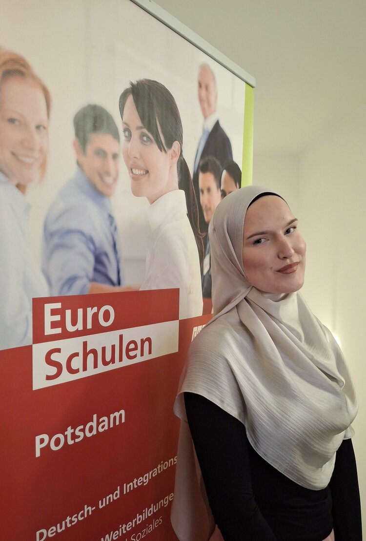 Eine Frau mit hellgrauem Kopftuch und schwarzem Oberteil lehnt sich leicht an einen Werbeaufsteller der 'Euro Schulen' in Potsdam. Sie lächelt sanft in die Kamera. Der Aufsteller zeigt ein Bild von lächelnden Menschen in Business-Kleidung sowie Texte zu den angebotenen Bildungsprogrammen, darunter Deutsch- und Integrationskurse sowie Weiterbildungen im sozialen Bereich. Der Hintergrund ist eine helle, neutrale Innenraumumgebung.