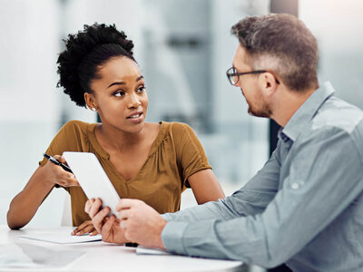 Eine Frau und ein Mann sitzen an einem Tisch und diskutieren über ein Dokument. Die Frau hält ein Tablet in der Hand und scheint etwas zu erklären, während der Mann aufmerksam zuhört. Die Szene deutet auf eine professionelle oder beratende Situation hin.