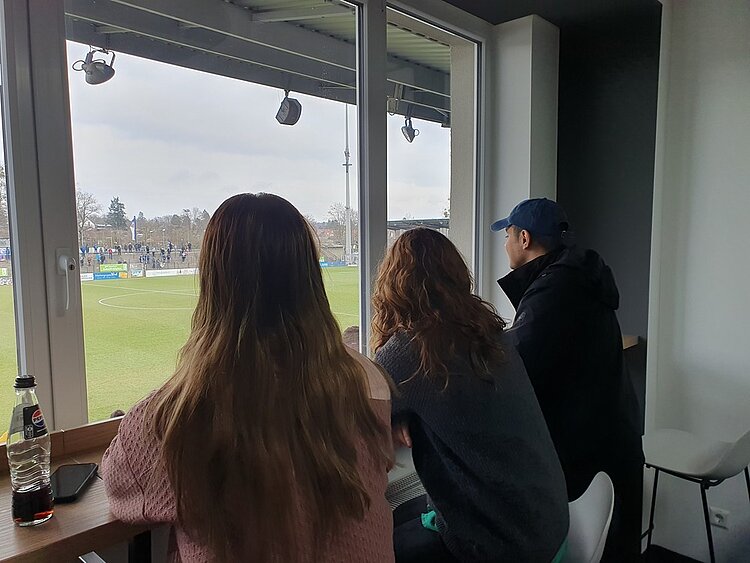 Stadionerlebnis der Extraklasse bei Turbine Potsdam Zwei Frauen und ein Mann schauen aus der VIP-Lounge durch ein großes Fenster auf das Geschehen auf dem Spielfeld.