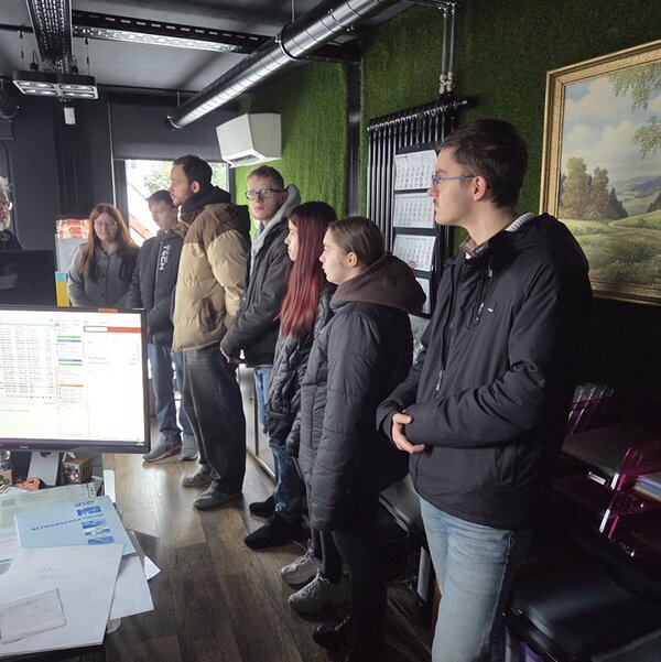 Mehrere Auszubildende der Euro-Schulen Hoyerswerda stehen in winterlicher Kleidung in einem modernen Büroraum einer Werbeagentur. Im Vordergrund befindet sich ein Schreibtisch mit Computerbildschirm, Tastatur, Papieren und Büromaterialien. Die Wände sind teilweise mit grünem Kunstrasen verkleidet, an einer Wand hängt ein gerahmtes Landschaftsbild. Im Hintergrund sind Regale mit Aktenordnern und eine Klimaanlage zu sehen. Die Auszubildenden stehen nebeneinander und blicken in Richtung einer Person oder eines Objekts außerhalb des Bildausschnitts. Die Szene vermittelt den Eindruck eines Gruppenbesuchs oder einer Führung durch die Agentur.