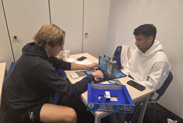 Zwei junge Männer spielen Schiffe versenken an einem Tisch. Einer setzt gerade Schiffe auf dem Spielfeld, während der andere konzentriert zuschaut.