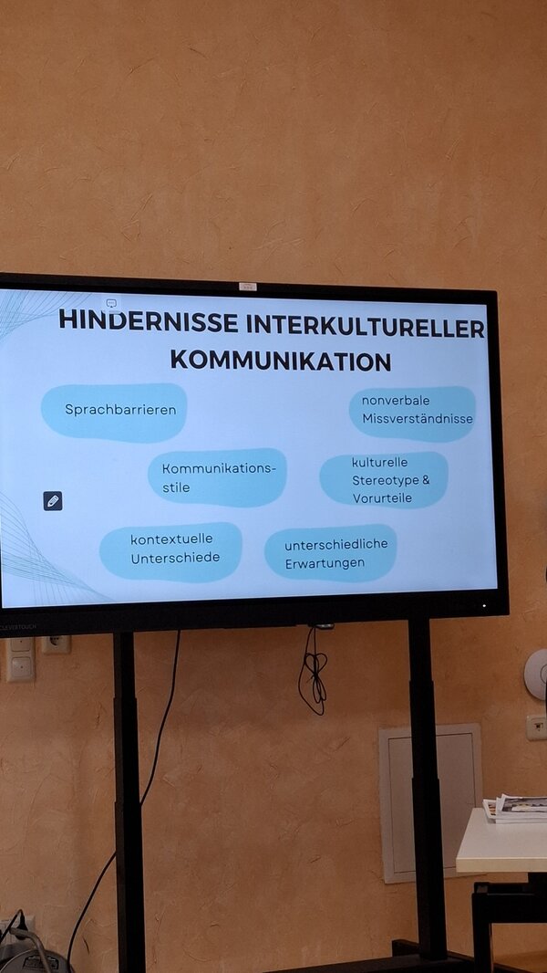 Ein großer Bildschirm zeigt eine Präsentationsfolie mit dem Titel 'Hindernisse interkultureller Kommunikation'. Darunter sind sechs hellblaue, unregelmäßig geformte Textfelder verteilt, die folgende Begriffe enthalten: 'Sprachbarrieren', 'nonverbale Missverständnisse', 'Kommunikationsstile', 'kulturelle Stereotype & Vorurteile', 'kontextuelle Unterschiede' und 'unterschiedliche Erwartungen'. Der Bildschirm steht vor einer orangefarbenen Wand, und darunter sind Kabel sichtbar, die zum Bildschirm führen. Rechts neben dem Bildschirm steht ein kleiner Tisch mit einigen Gegenständen darauf.