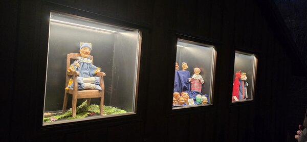Beleuchtete Schaufenster mit traditionellen Handpuppen und Figuren in einer dunklen Außenwand, nachts aufgenommen.