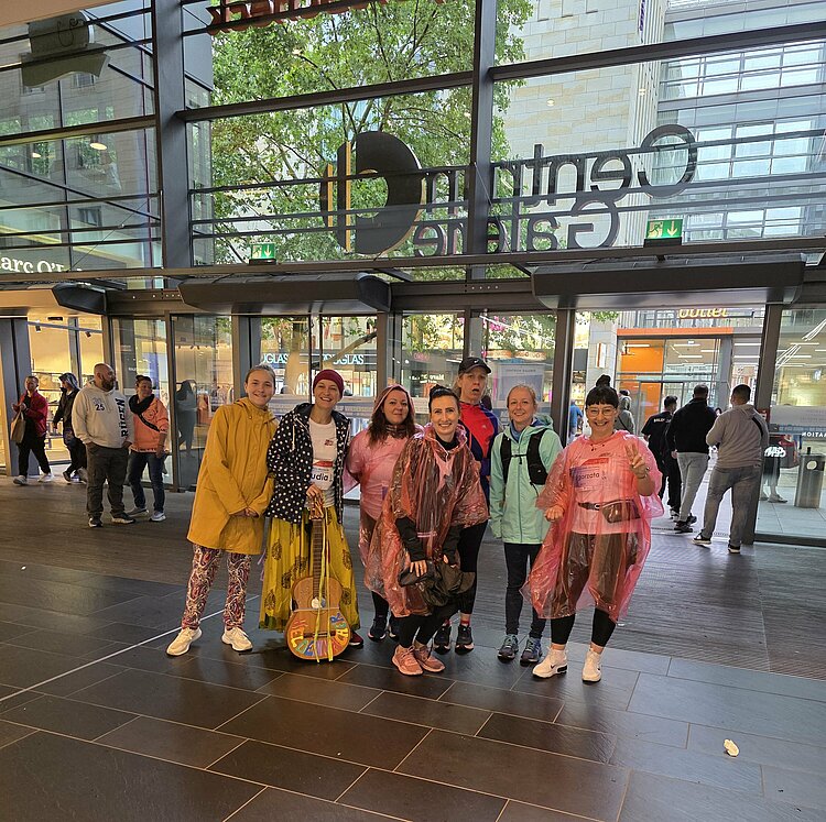 Euro-Schulen & Euro Akademie bei der Rewe Team Challenge 2025 Eine Gruppe von acht Personen in Laufkleidung posiert im Eingangsbereich der Centrum Galerie. Einige tragen bunte Regenponchos, eine Gitarre steht in der Mitte.
