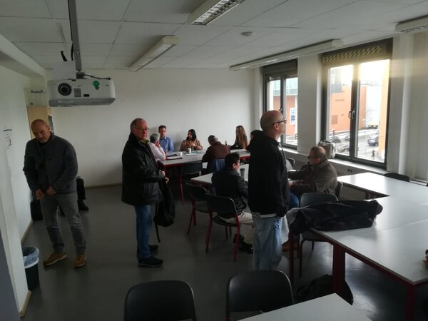 Mehrere Personen in einem Seminarraum sitzen und stehen an Tischen; rechts große Fenster, oben ein Deckenbeamer.