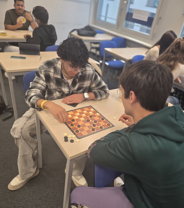 Zwei junge Männer sitzen an einem Tisch und spielen Dame. Beide konzentrieren sich auf das Spielbrett, während im Hintergrund weitere Personen sitzen und sich unterhalten.