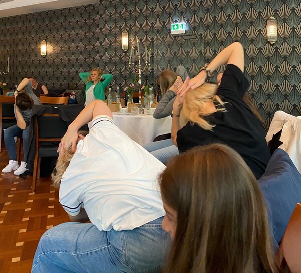 Eine Gruppe von Menschen sitzt auf Stühlen in einem Raum mit dekorativen Wandmustern und führt eine Übung durch, bei der sie ihre Arme hinter dem Kopf kreuzen.