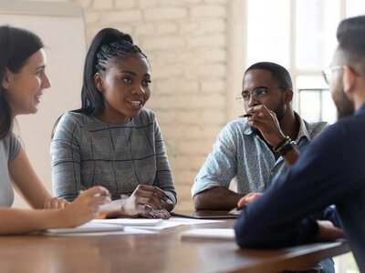 Vier Personen sitzen an einem Konferenztisch in einem hellen Raum. Die Frau in der Mitte, mit geflochtenem Haar und einem karierten Oberteil, lächelt und spricht mit den anderen. Links von ihr sitzt eine Frau mit dunklen Haaren, rechts ein Mann mit Brille und einem gepunkteten Hemd, und ein weiterer Mann ist teilweise rechts im Bild zu sehen.