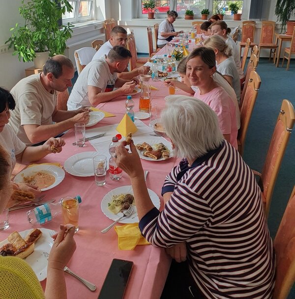 Die Teilnehmerinnen und Teilnehmer sitzen gemeinsam an der langen Essenstafel und genießen ihre selbst zubereiteten Speisen. 