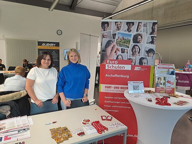 Frau Beller und Frau May strahlen am Messestand neben dem Roll-up der Euro-Schulen um die Wette in die Kamera. Auch einige Messegeschenke sind auf den Tischen zu sehen.