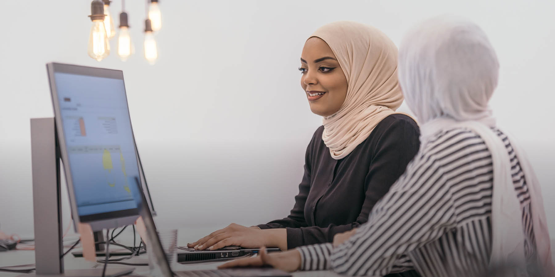 Zwei Frauen sitzen vor einem Computer in einer modernen Arbeitsumgebung.