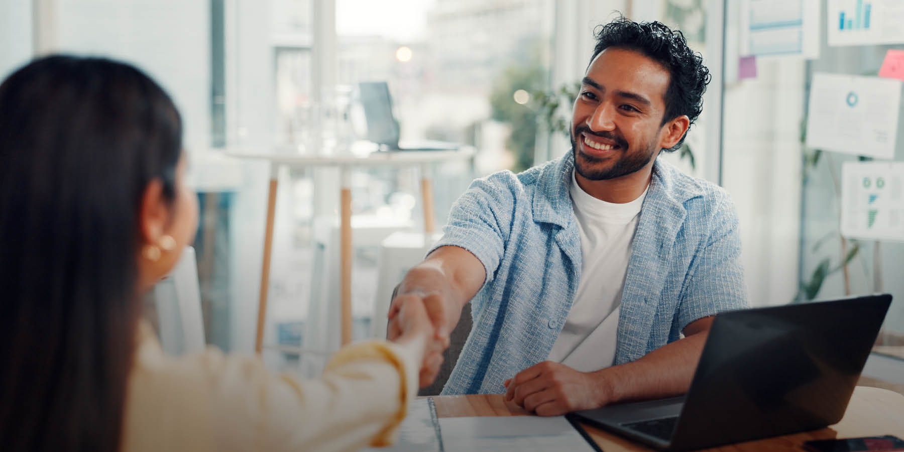Ein Mann sitzt vor einem Laptop in einer modernen Arbeitsumgebung. Er schüttelt einer Frau lächelnd die Hand.
