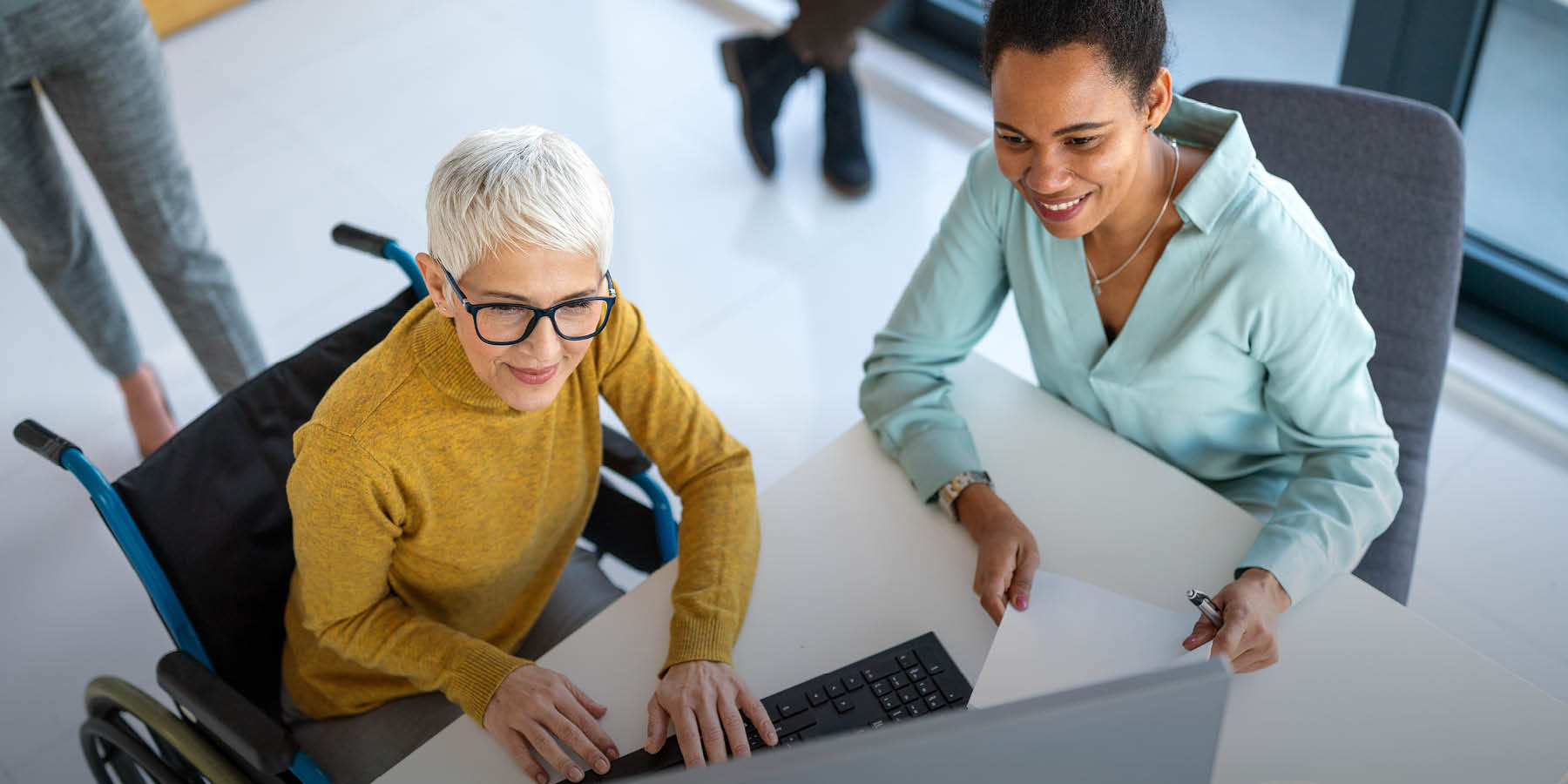 Das Bild zeigt eine ältere Frau im Rollstuhl und eine jüngere Frau von oben. Die beiden sitzen gemeinsam an einem Tisch und arbeiten am Computer.