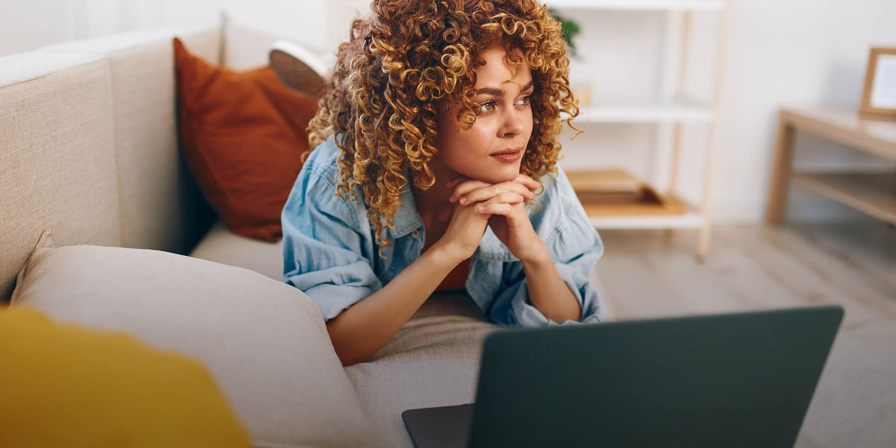 Eine Frau liegt gedankenverloren auf dem Sofa, den Kopf auf die Hände gestützt. Vor ihr steht ein Laptop.