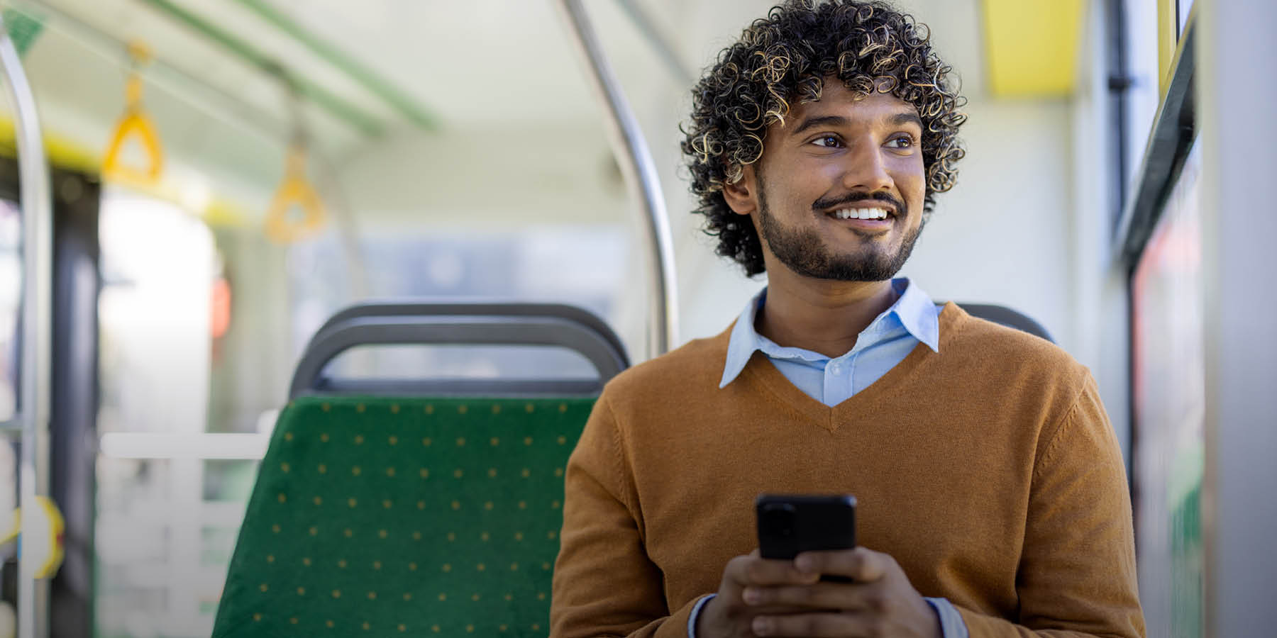 Ein Mann sitzt im Bus. Er hat ein Handy in der Hand und sieht lächelnd aus dem Fenster.