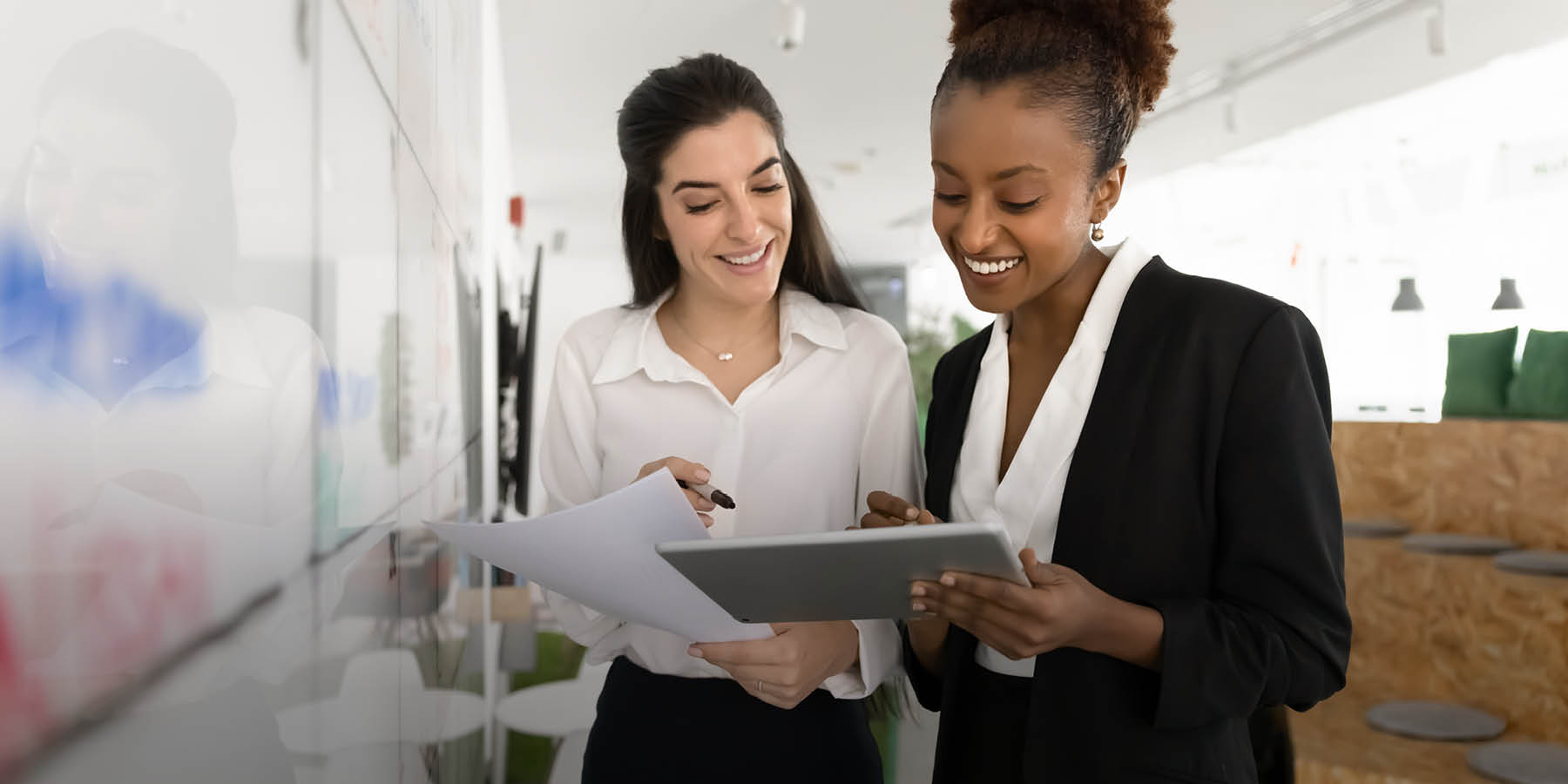 Zwei Frauen in Brüokleidung stehen neben einem Whiteboard und blicken lächelnd auf das Tablett, das die linke Frau in den Händen hält.