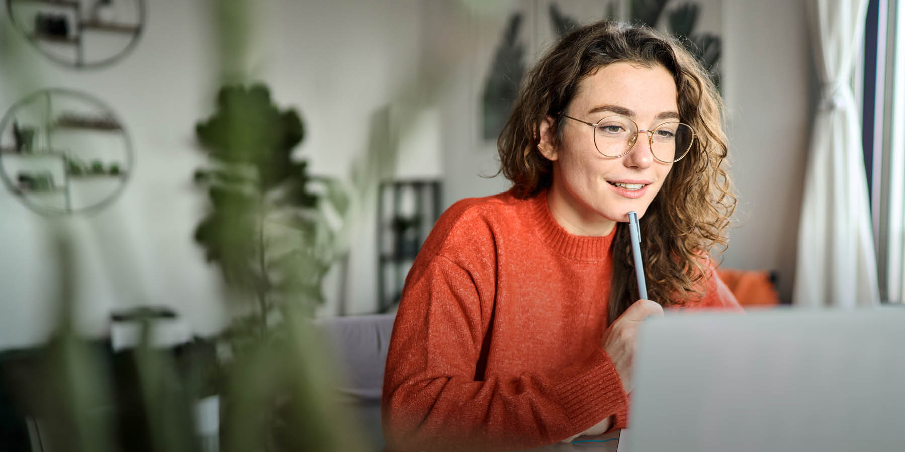 Eine Frau sitzt zu Hause vor einem Laptop und blickt auf den Bildschirm. Sie hat das Kinn auf einen Stift gestützt.