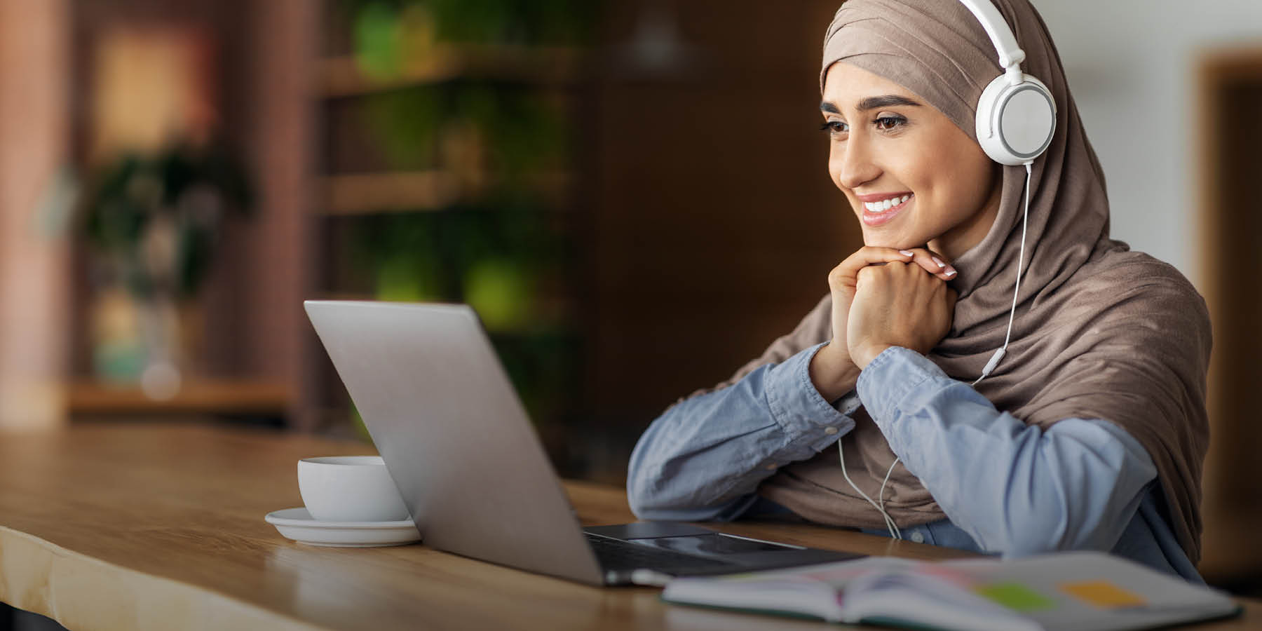 Eine Frau sitzt vor einem Laptop und blickt lächelnd auf den Bildschirm. Sie trägt große Kopfhörer.